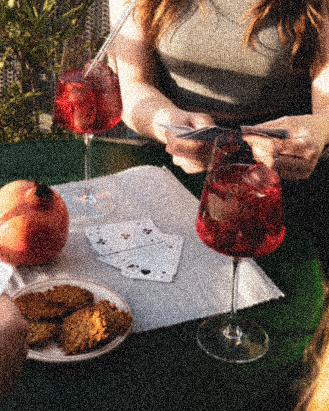 Eine Frau die Spielkarten in der Hand hat und ein Tisch mit GRANADA NIGHTS Spritz, einem Granatapfel und einem Teller mit Snacks.