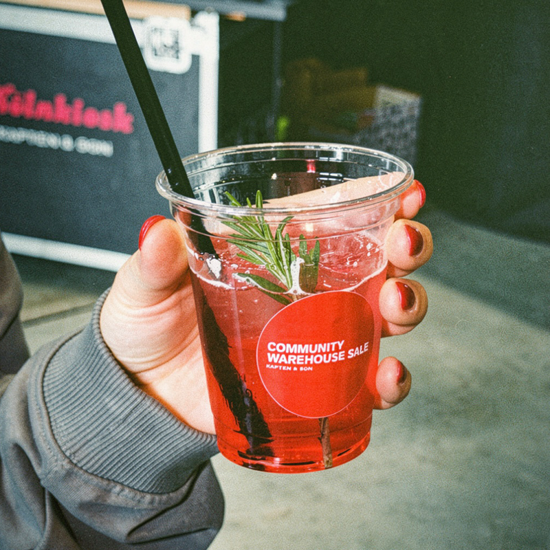 Ein Becher GRANADA NIGHTS Spritz mit Strohhalm, Rosmarin und einem Aufkleber mit der Aufschrift "Community Warehouse Sale Kapten & Son".